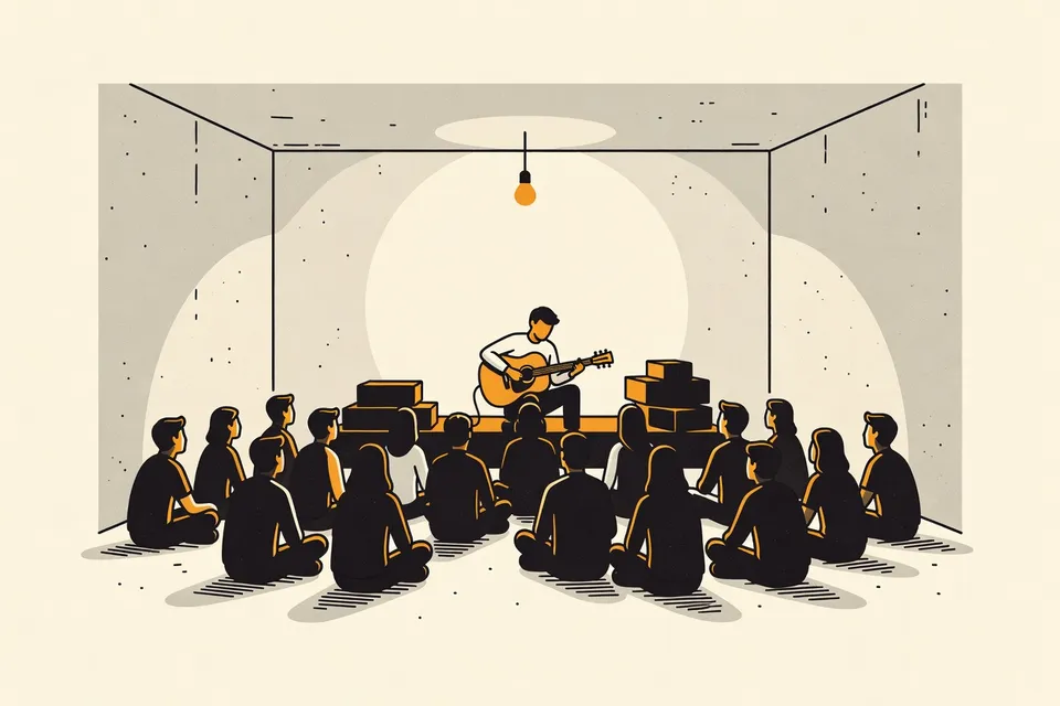 A wide shot of a dim concrete room with a small audience sitting on the floor, facing a stage with a musician tuning a guitar. The scene is intimate and quiet.