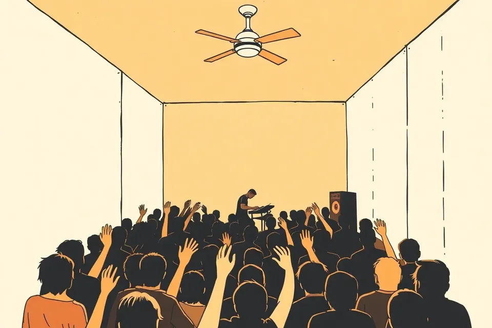 A dim, intimate live music venue with a small crowd of silhouetted figures facing a stage where a musician plays a synthesizer, under a single ceiling fan and bare concrete walls.
